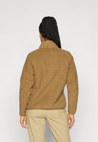 Pullover in pile marrone con finitura testurizzata, colletto alto e maniche lunghe. Indossato con pantaloni beige, mostrando una vestibilità e un taglio rilassati.