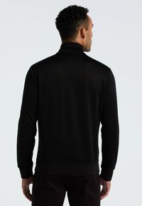 Zwarte ritsjack met hoge kraag en geribde manchetten. Beschikt over textuurstof en contrasterende teal accenten aan de zijkanten.