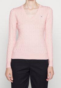 Maglione rosa a lavorazione a treccia con scollatura a V, maniche lunghe, polsini e orlo a coste. Presenta un piccolo logo ricamato sul petto.