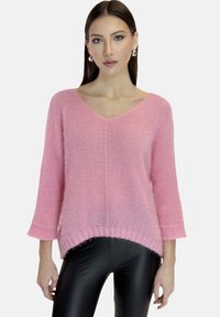 Rosa gestrickter Pullover mit V-Ausschnitt, Dreiviertelärmel und einem längeren Rückensaum. Weiche Textur und gerippte Bündchen. Kombiniert mit schwarzen Leggings.
