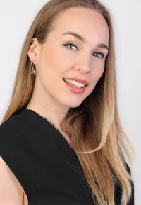 Gouden creolen met bladvormen, gedragen door een vrouw met lang, steil lichtbruin haar, in een zwarte top met rafelige randen.