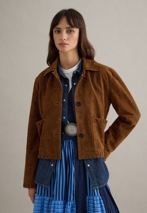 Junge Frau mit brauner Wildlederjacke über einem mehrlagigen, blau gestreiften Kleid und Jeanshemd, steht mit einer Hand in der Hüfte.