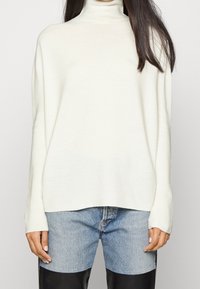 Kremowy sweter z golfem, długimi rękawami, miękką teksturą, luźnym krojem oraz prążkowanymi detalami przy dekolcie i u dołu. W zestawie z jasnym dżinsami.