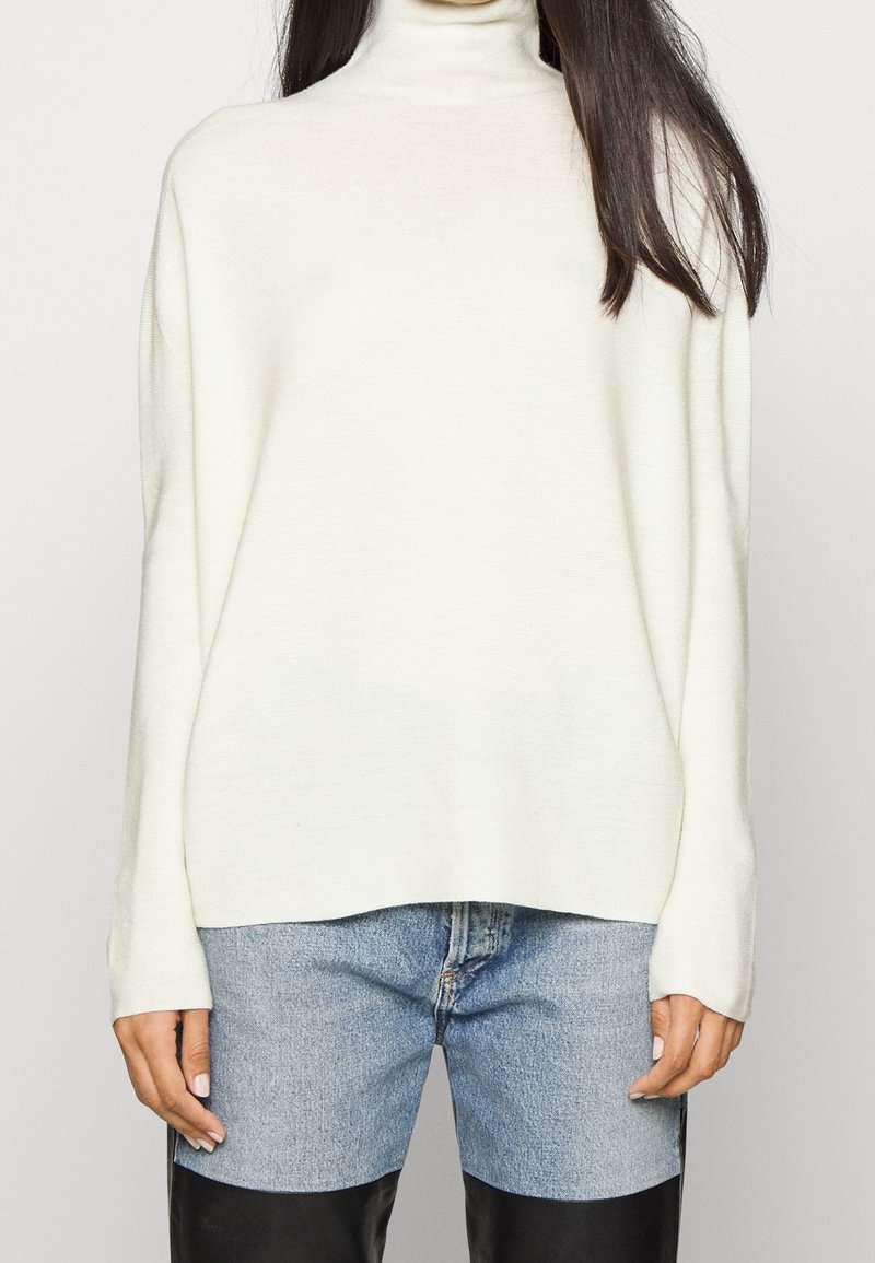 Kremowy sweter z golfem, długimi rękawami, miękką teksturą, luźnym krojem oraz prążkowanymi detalami przy dekolcie i u dołu. W zestawie z jasnym dżinsami.