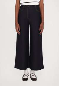 Bredde ben mørke denim bukser med oransje søm, høy midje, frontlommer og knappelukking, kombinert med hvite sokker og mørke sko.