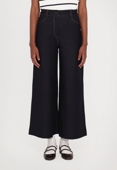 Wide-leg dark denim trousers with orange stitching, high waist, front pockets, and a button closure, paired with white socks and dark shoes.