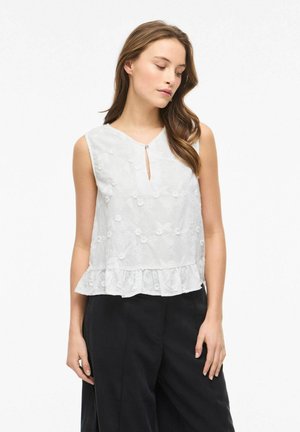 Kobieta ubrana w białą, bezrękawnikową, kwiatowo haftowaną bluzkę z falbanistym dołem i czarne spodnie, patrzy w dół w lewo.