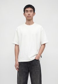 Junger Mann mit kurzen schwarzen Haaren, der ein schlichtes weißes kurzärmeliges T-Shirt und dunkle Jeans trägt, steht vor einem hellgrauen Hintergrund.