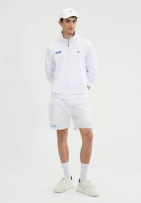 Chaqueta deportiva blanca con cremallera y pantalones cortos, con logotipos de la marca en azul. Combinada con una gorra blanca a juego y zapatillas blancas. Textura suave.