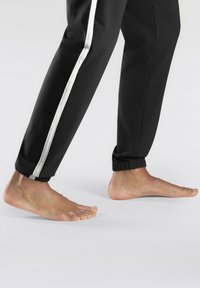 Schwarze Jogginghosen mit einem weißen Seitenstreifen, ausgestattet mit einem elastischen Bündchen am Knöchel. Ein nackter Fuß ist neben dem Hosenbein zu sehen.