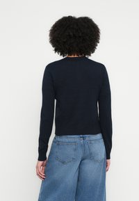 Tumši zils džemperis ar adījumu, garām piedurknēm, apaļu kaklu un ribotām apmalēm, kombinēts ar gaismas zilu platām džinsa biksēm ar aizmugurējām kabatām.