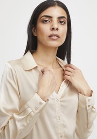 Beige Satinbluse mit klassischem Kragen, Knopfleiste vorne und langen Ärmeln. Sie hat einen dezenten Glanz und vier Knöpfe an jedem Ärmelbündchen.