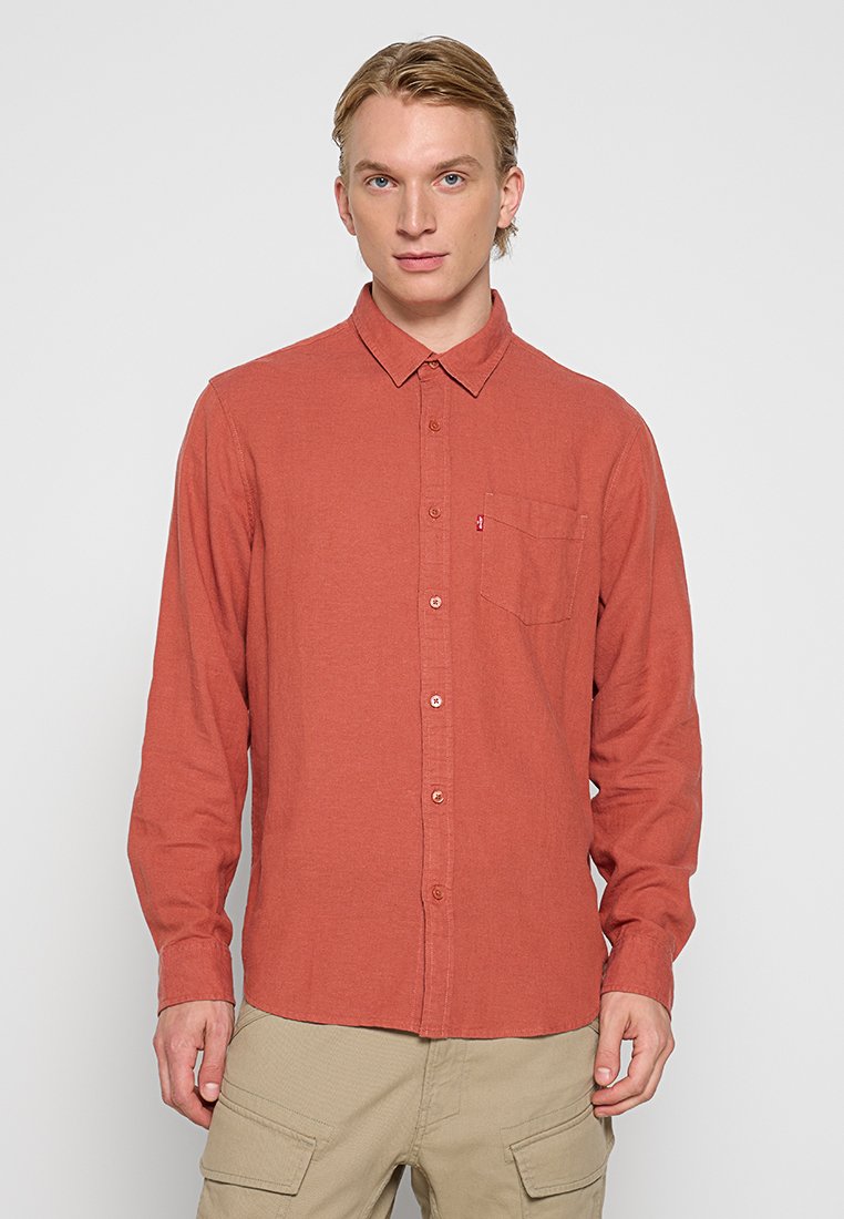 Chemise à col, à manches longues de couleur rouille, avec une poche poitrine unique arborant une petite étiquette de logo. Texture de tissu légère.