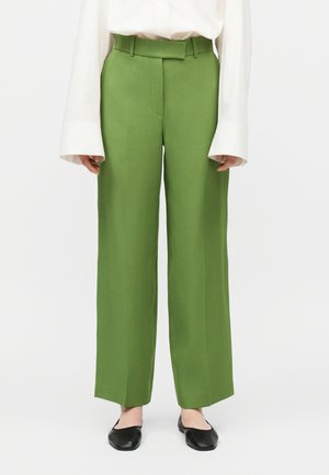 Zeleno hlače z visokim pasom in ravnimi hlačnicami s pasom v enaki barvi, kombinirane z belo bluzo z dolgimi rokavi in črnimi ravnimi čevlji.