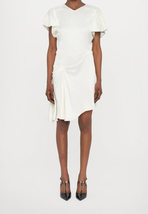 Femme portant une robe blanche mi-longue avec des manches volantées et un ourlet asymétrique, associée à des talons noirs à brides en T et bouts pointus.