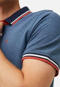 Niebieska koszulka polo z teksturowanej tkaniny, z granatowym kołnierzykiem i czerwonym paskiem akcentującym, oraz kontrastującymi paskowanymi mankietami.