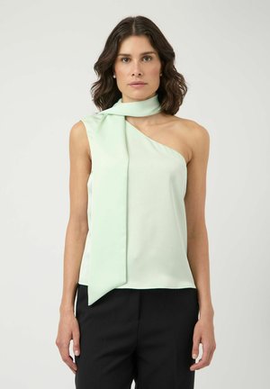 Femme portant un top asymétrique vert pâle sans manches avec un long détail de foulard sur une épaule, associé à un pantalon noir, debout sur un fond blanc.