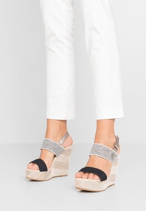 Osoba ubrana w białe spodnie i beżowe sandały na koturnie z czarnymi i szarymi paskami, stojąca na jasnoszarej podłodze na tle białej ściany.