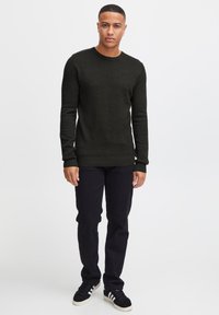 Maglione lavorato a maglia verde scuro con maniche lunghe, scollatura rotonda, orlo a costine, abbinato a pantaloni neri e sneakers nere.