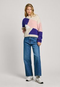 Un pull en tricot coloré avec de grandes formes géométriques en crème, rose, violet et blanc. Porté avec un jean bleu taille haute et des baskets.
