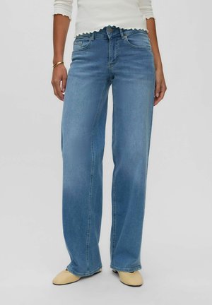 Femme portant un jean bleu clair à jambes larges, des chaussures beiges, un haut blanc à manches longues et un bracelet en or au poignet gauche, debout devant un fond uni.
