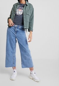 Chaqueta verde corta, camiseta gráfica gris, jeans de pierna ancha azul claro, zapatillas blancas con acentos sutiles y calcetines blancos con detalles rojos.