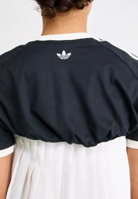Juodas medvilninis marškinėlis su baltu banguotu apykakle ir rankogaliais, apatinėje dalyje turi klostuotą baltą audinį ir mažą baltą logotipą nugaros pusėje.