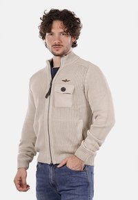 Maglione beige lavorato a maglia con zip, con un motivo testurizzato, colletto alto, tasca sul petto e un emblema sulla spalla. Abbinato a jeans blu.