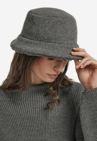 Szary filcowy kapelusz typu bucket z szerokim, postrzępionym rondem. Teksturowana powierzchnia i zaokrąglony kształt uzupełniają prążkowany szary sweter noszony pod spodem.
