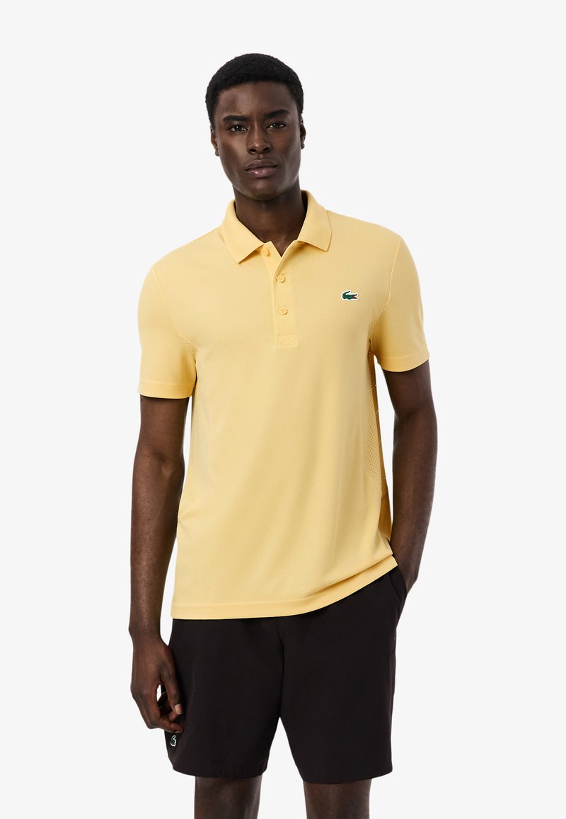 Joven de pie, vistiendo una camiseta tipo polo amarilla clara con un logo en el pecho y pantalones cortos negros, sobre un fondo blanco liso.