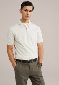 Off-white polo s srajco z zadrgo pri ovratniku, v kombinaciji z olivno zelenimi hlačami in rjavim pasom, ki prikazuje strukturirano tkanino.