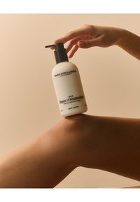 Bouteille cylindrique blanche avec un couvercle à pompe noir, étiquetée "N°12 Objets d'Amsterdam," reposant sur un genou bronzé avec une texture de peau lisse.