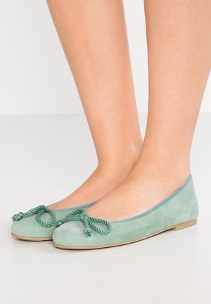 Pieds portant des ballerines en daim vert clair avec un nœud torsadé en corde sur un fond clair.