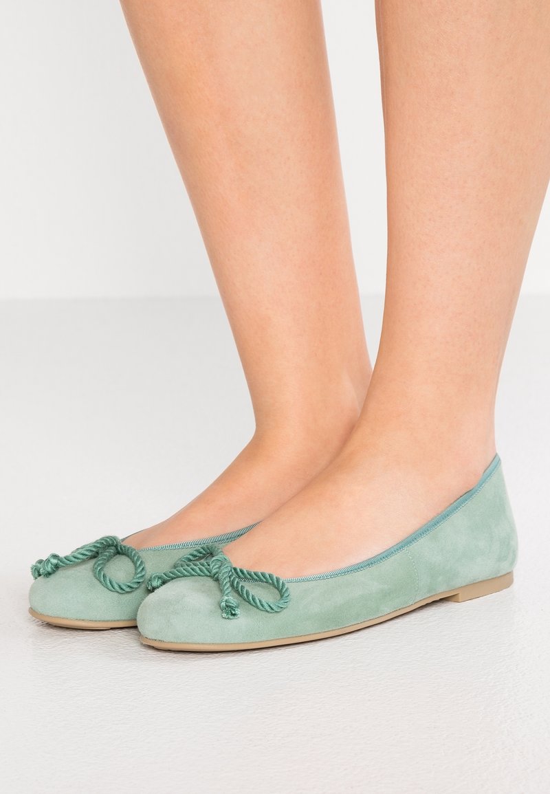 Pieds portant des ballerines en daim vert clair avec un nœud torsadé en corde sur un fond clair.