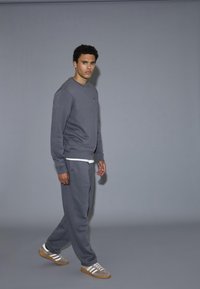 Grå sweatshirt med rund halsringning och ribbade muddar, matchad med grå sweatpants. Modellen står framför en grå bakgrund.