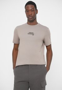 Young man with dark skin wears a beige Armani Exchange T-shirt and gray cargo pants, standing with hands in pockets against a plain background.