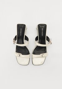 Sandalias de tacón de charol blanco con espalda abierta y dos correas; punta cuadrada; detalle de hebilla ajustable; forro interior negro.