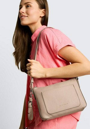 Jonge vrouw in een roze shirt die een beige leren schoudertas met textuur en verstelbare band vasthoudt tegen een effen achtergrond.