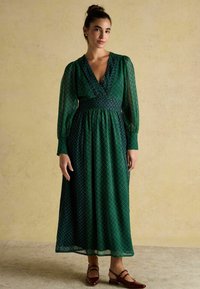 Duga zielona sukienka w geometryczny wzór, z głębokim dekoltem w kształcie litery V, marszczonym pasem i przezroczystymi długimi rękawami. Noszona z burgundowymi balerinami.