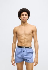 Lys blå boxershorts av bomull med en svart midjebånd som har "BOSS"-logoen. Knappelukking foran, glatt tekstur.