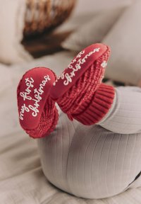 Czerwone dziergane buciki dziecięce z napisem "moje pierwsze Święta", wyposażone w gładką czerwoną podeszwę i prążkowany mankiet na kostce dla ciepła. Widoczne szare prążkowane legginsy.