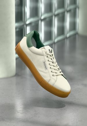 Weißer Ledersneaker mit grünem Fersenlaschen, brauner Sohle und "BOGNER"-Logo an der Seite, schwebt über einem grauen Boden nahe Glasbausteinen.