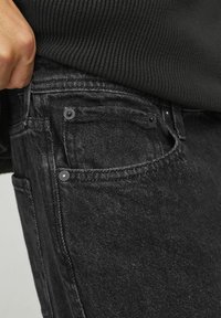 Jeans de mezclilla negra con una textura desteñida, que cuentan con un bolsillo delantero con remaches de metal y un ajuste ceñido, parcialmente cubiertos por un suéter oscuro.