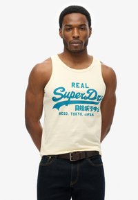 Kremowy top na ramiączkach z tealowym napisem "SuperDry", okrągłym dekoltem i luźnym krojem. Noszony z ciemnoniebieskimi dżinsami i brązowym paskiem.