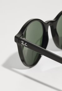 Ochelari de soare Ray-Ban negri cu lentile verzi, formă rotundă, rame din plastic lucios și logo argintiu pe braț.