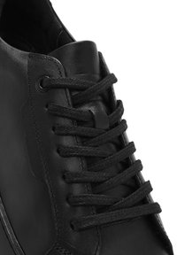 Sneaker en cuir noir avec bout rond, surface texturée et lacets noirs proéminents passés à travers des œillets métalliques. Design minimaliste.