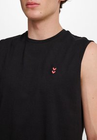 Man med en ärmlös svart tröja med en liten rosa dubbel chevron-logotyp på vänster bröst mot en enkel vit bakgrund.