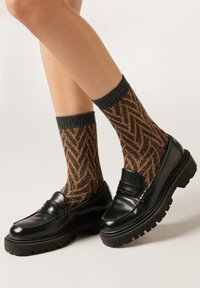 Schwarze Leder-Loafer mit dicker Sohle, kombiniert mit grauen und orangefarbenen Socken im Fischgrätmuster. Glatte Struktur und markante Designdetails.