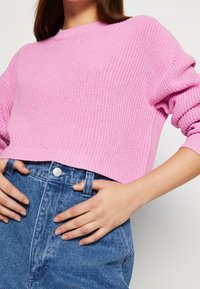 Rosa gestrickter Pullover mit rundem Ausschnitt und senkrecht geschnittenen Schultern, kombiniert mit hochgeschnittenen blauen Jeans.