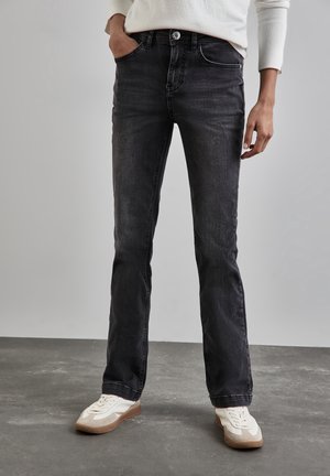 Underkroppen af en person iført falmede sorte jeans med lige ben, hvide sneakers og en hvid langærmet top, stående på et gråt gulv.